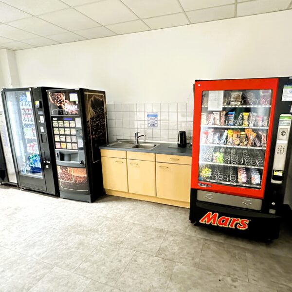 Foto vom Aufenthaltsraum für Teilnehmende. Zu sehen sind verschiedene Snackautomaten und ein Waschbecken.