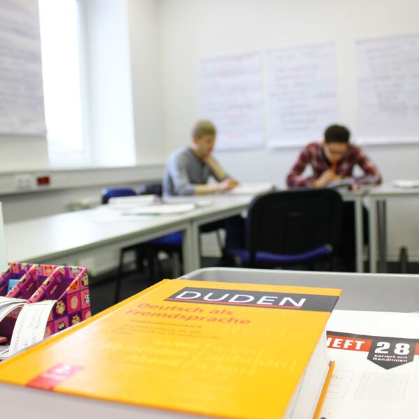 Foto der German Language Academy am Bahnhof. Zu sehen ist eine Lernsituation mit Personen. Ein Duden liegt auf einem Tisch.